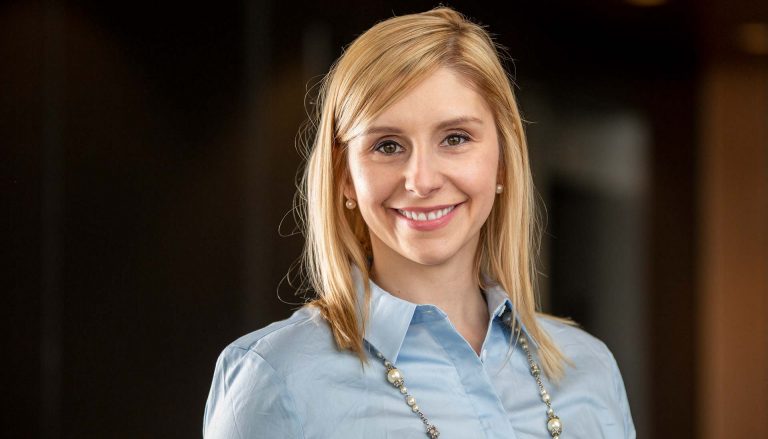 Young, smiling woman with long, blonde hair, a blue shirt, and a silver and pearl necklace.