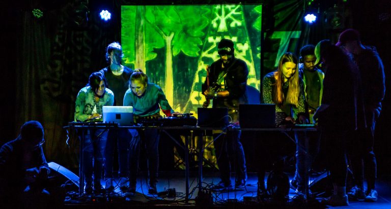A group of people making music on a stage with computers and with blue and green lights projected on to them.