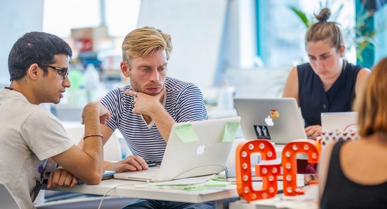 Young people in an open office environment, looking at laptops and collaborating.