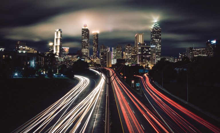 Dark time lapse of highway leading to lit cityscape, with red lines from car lights