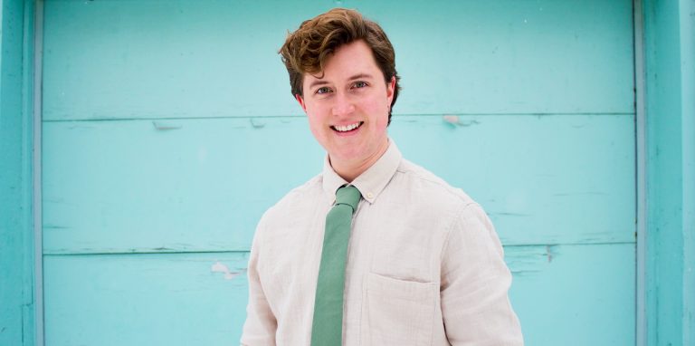 Young smiling man with curly, short hair, in a dress shirt and green tie, standing in front of a cyan-coloured wall.