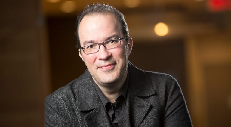 A smiling man with short, thinning hair, glasses and a dark-grey shirt and jacket.