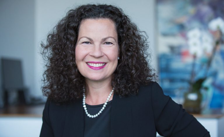 Smiling woman with long, dark, curly hair, pink lipstick, a pearl necklace and a black shirt and jacket.