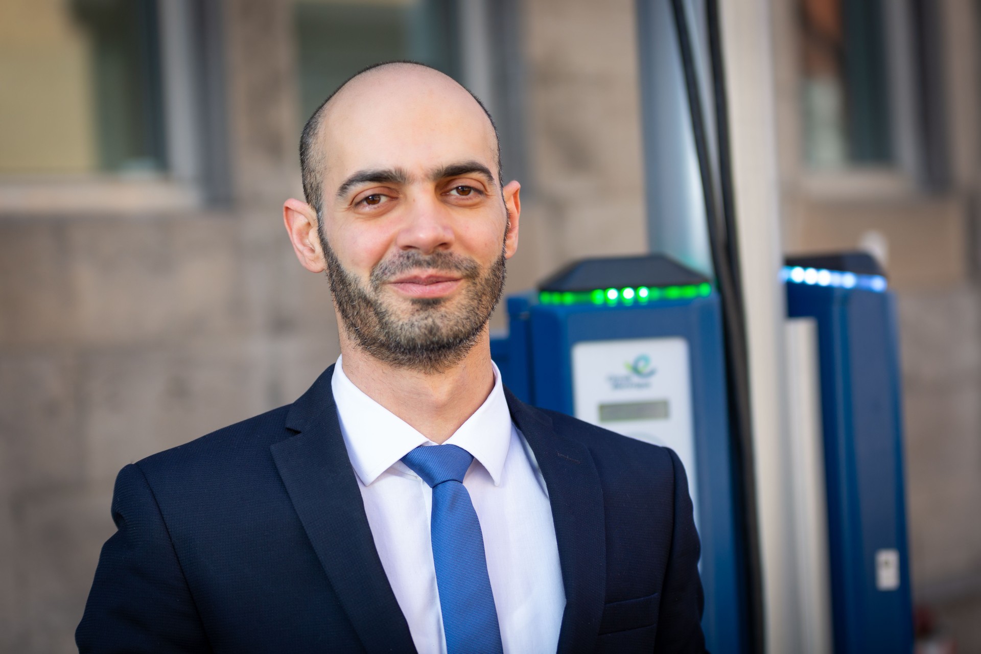 Man with short beard in blue blazer and tie standing in front of EV charging station