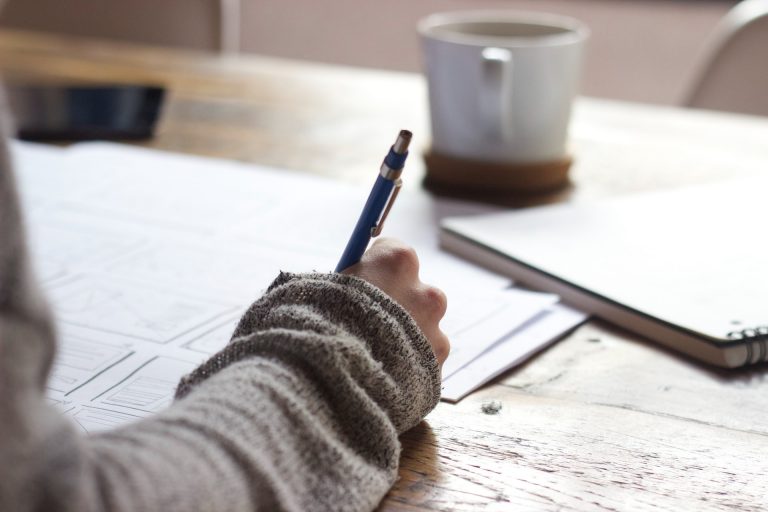 Hand holding pen and writing notes with white papers spread out on desk.