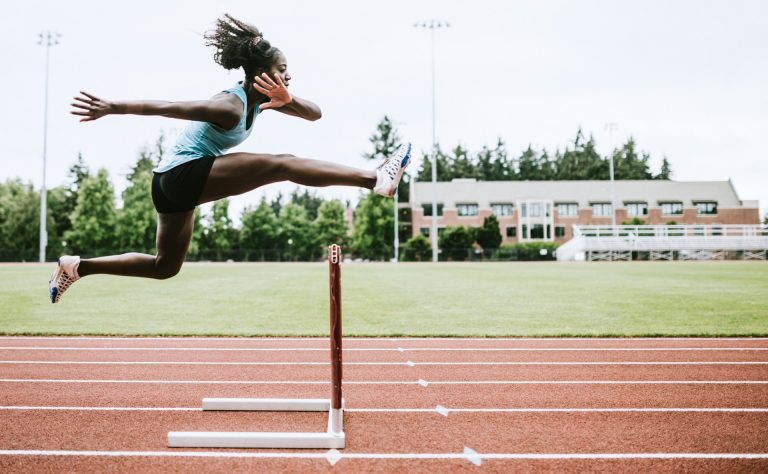 A young, female, Black athlete jumping over a hurdle on a race track.