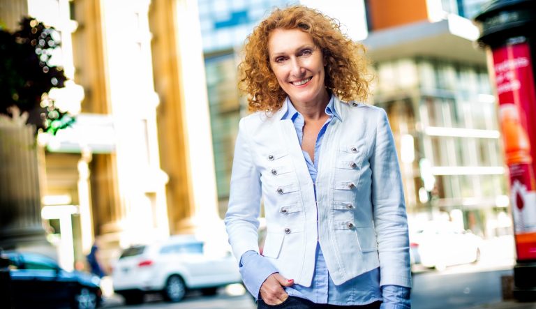 Woman with long, curly red hair, jeans, a blue shirt and white jacket standing on a city street.