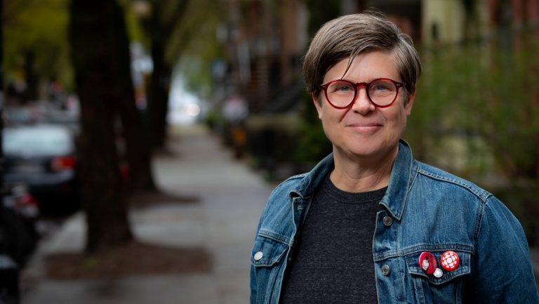 Woman with short hair, glasses and jean jacket standing on a sidewalk outside