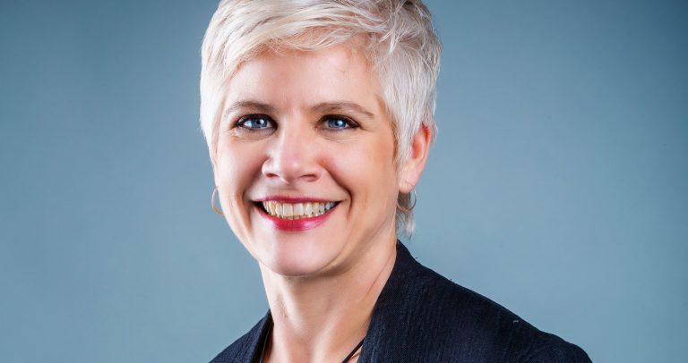 Woman with short, blonde hair, a dark jacket and a leather necklace.