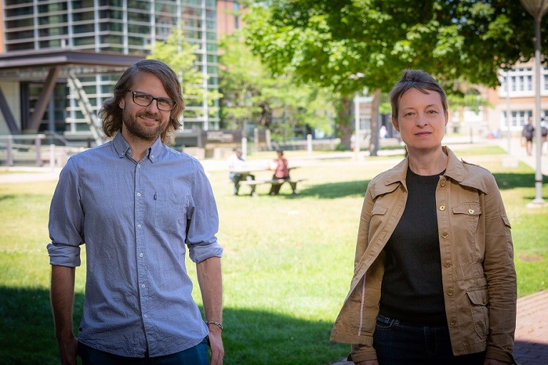 Man with glasses in blue collared shirt and woman in brown jacket standing near a lawn