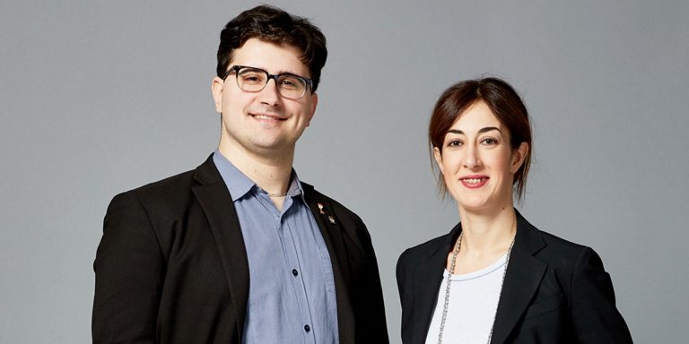 A smiling man and woman standing together against a grey background.