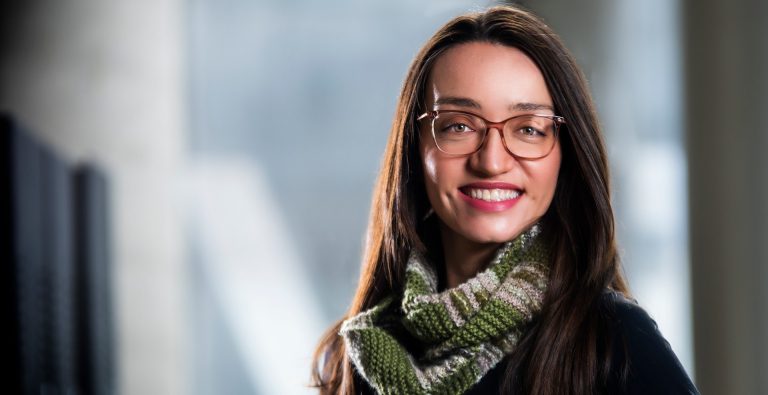 A young, smiling woman with long, dark hair, glasses, a green wool scarf and a black top.