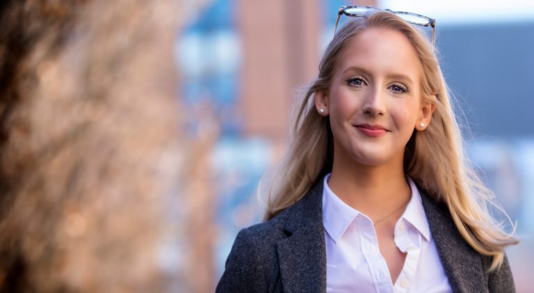 A young, smiling woman with long, blonde hair, glasses on her head, a white shirt and grey jacket.