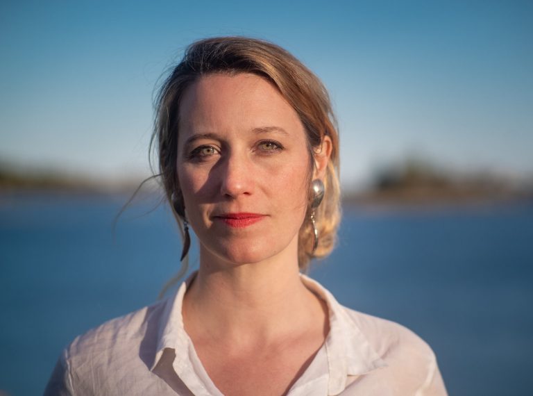 Blonde woman with hair tied back wearing white blouse, standing in front of blurry water backdrop