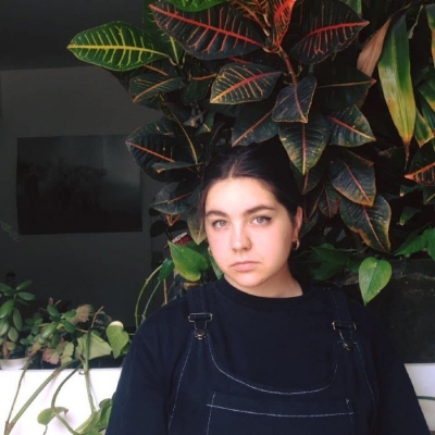 Woman with hair tied back wearing overalls sitting in front of tall leafy plant