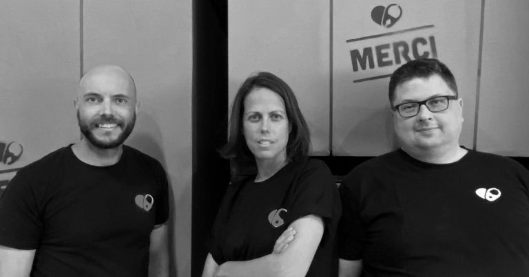 Two men and a woman with black T-shirts on, standing in front of large cardboard boxes, with the word "merci" written on them.