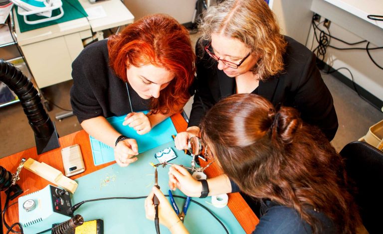 Three woman around a table, working on what looks like an electronic circuit.