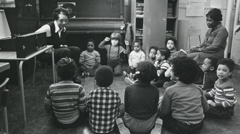 An archive image of a classroom with two teachers and several children sitting in a circle.
