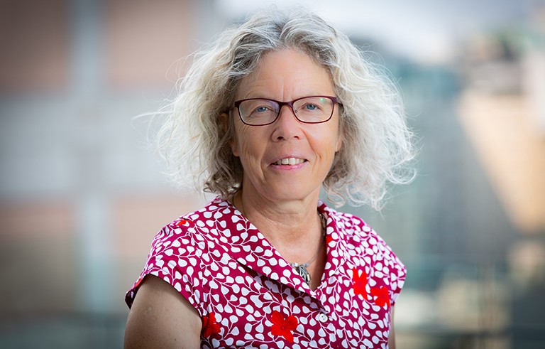 Smiling woman with shoulder-length blonde hair, glasses and a red and white patterned shirt.