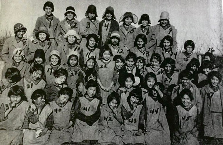 An archive photo of young Indigenous teenage girls at a residential school, each with a number on their pinafores.