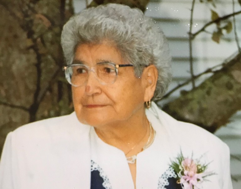 A smiling elderly woman with a white jacket, a printed shirt and a bouquet of pink flowers in her lapel