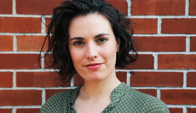 Young smiling woman with dark, shoulder-length curly hair and a green patterned shirt, standing in front of a brick wall.