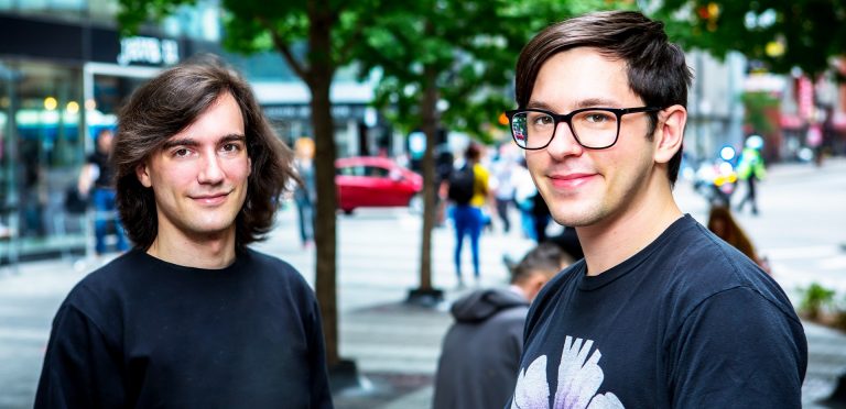 Two smiling young men standing in an urban setting.