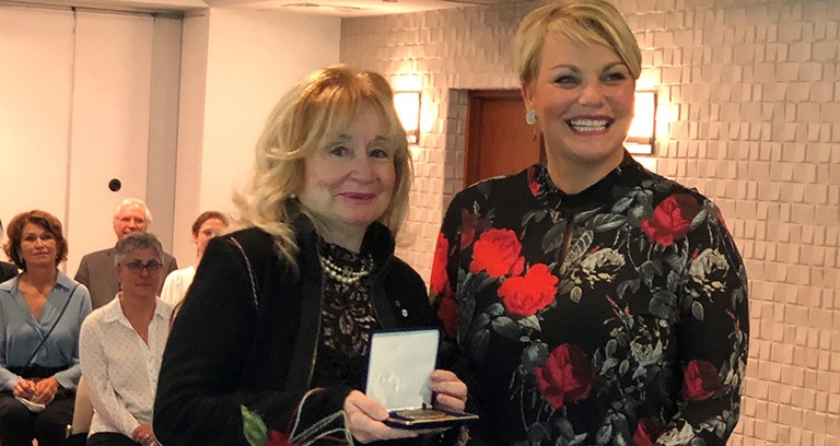 Two smiling women in a salle with seated people, and one woman holding an award.