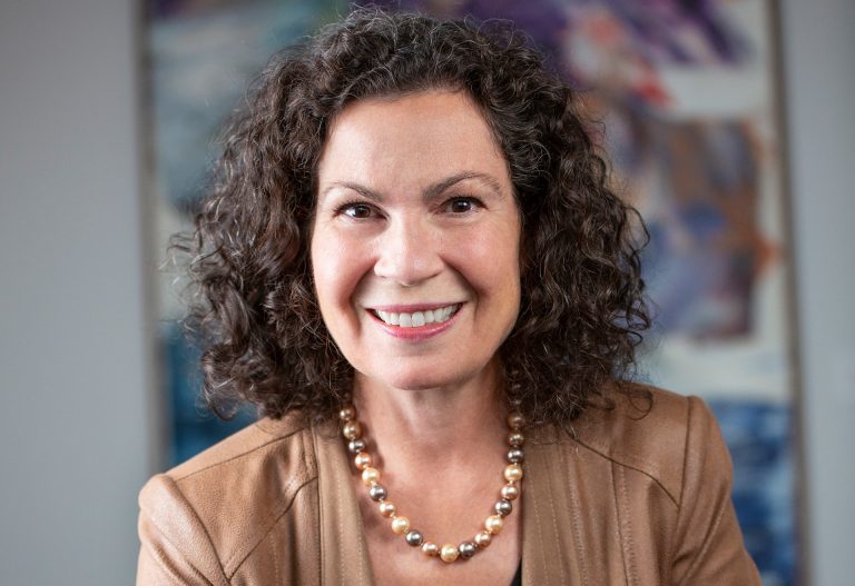 Smiling woman with shoulder-length dark, curly hair, a necklace, black top and tan leather jacket.