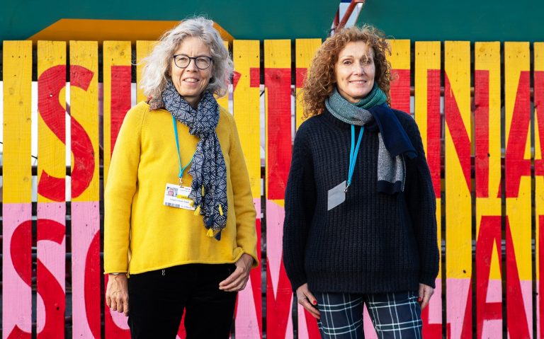 Two women, one with long, curly blonde hair and one with curly, red hair, standing in front of a colourful fence.