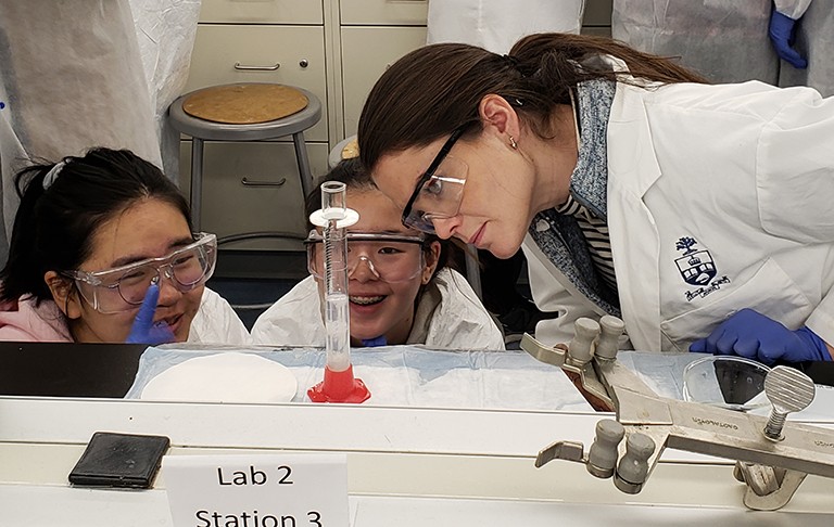 Two young girls and a woman in safety goggles watching a science experiment in a lab.