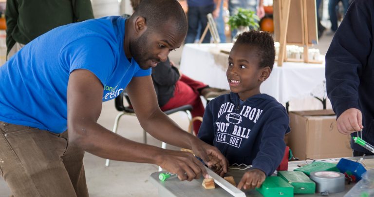 A Black man in a blue T-shirt explaining a science experiment to a young, smiling Black boy in a blue hoodie.