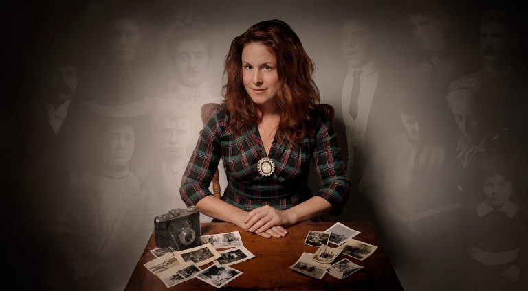 A woman with long, red hair, slightly smiling at the camera, with her hands folded on a table containing a camera and a number of photographs.