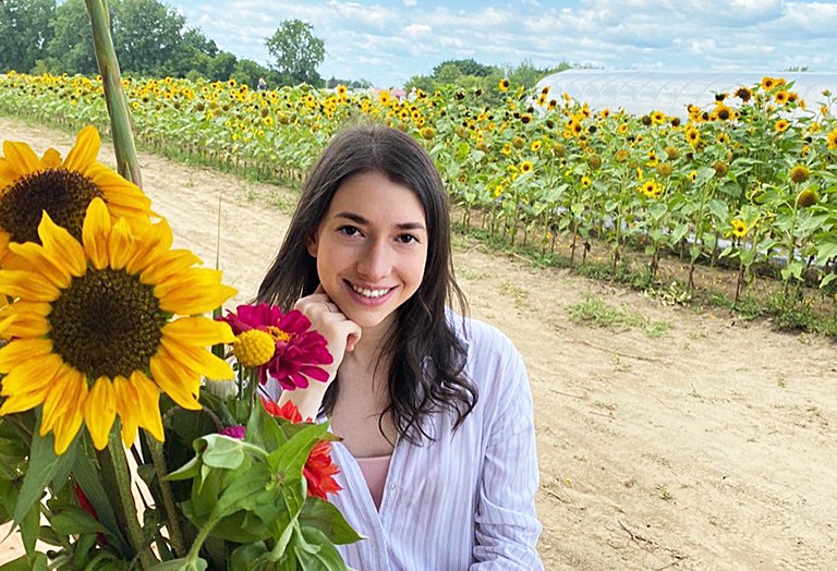 A smiling young woman with long, dark hair standing in a field of sunflowers.