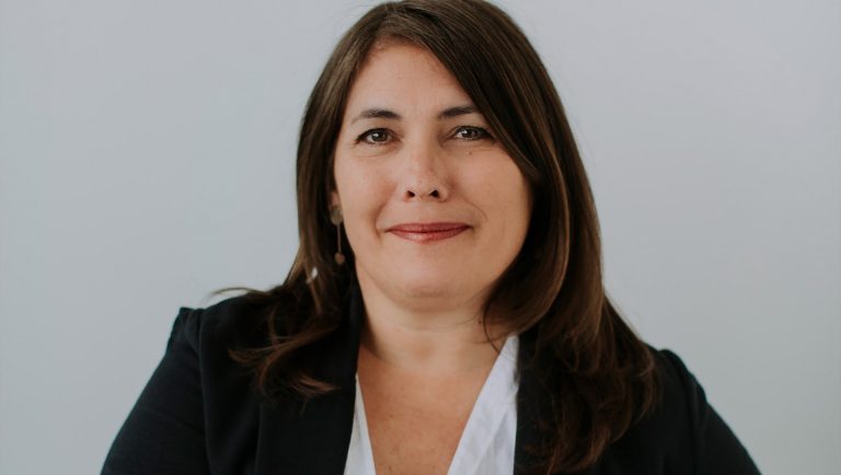 Smiling woman with long, dark hair, red lipstick and a white blouse under a black jacket.