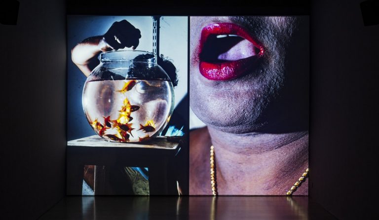 Image of a large mouth with bright crimson lipstick in the background, with a fishbowl filled with goldfish in the foreground.