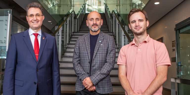 Three smiling men standing in a line holding their hands in front of them and standing before a staircase.