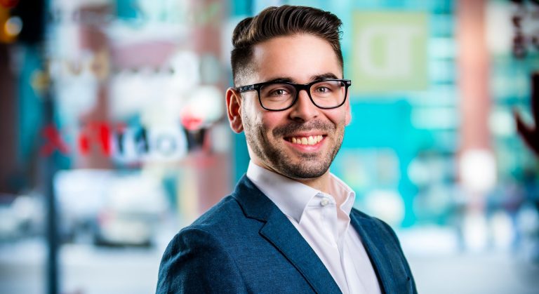 Young, smiling man with dark-rimmed glasses, a close-cropped beard and short hair, with a white shirt and blue dress jacket.