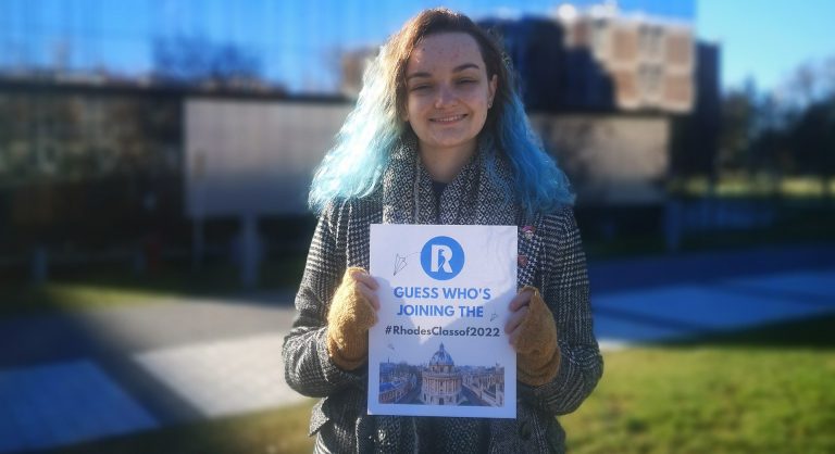 A smiling young woman with blonde and green hair, a winter coat and fingerless gloves, holding a small sign that says, "Guess who's joining the #RhodesClass2022."