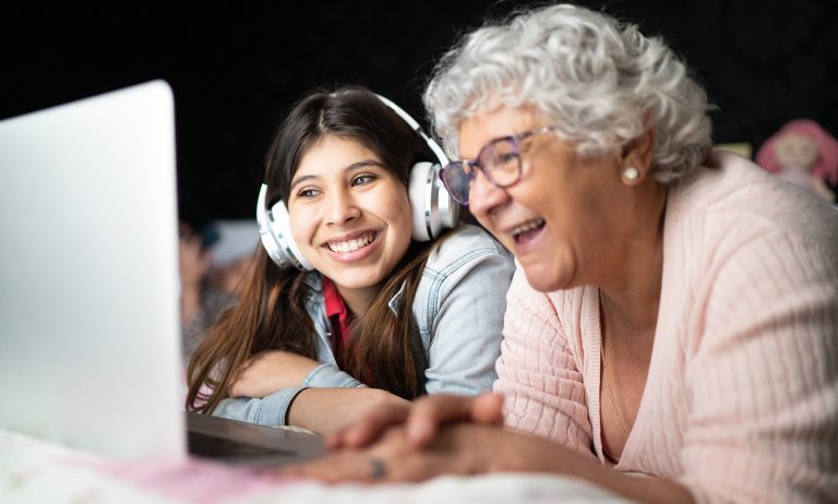 Pictured: A young smiling woman with headphones, and beside her, an older woman who is laughing. Both of them looking at the screen on a laptop.