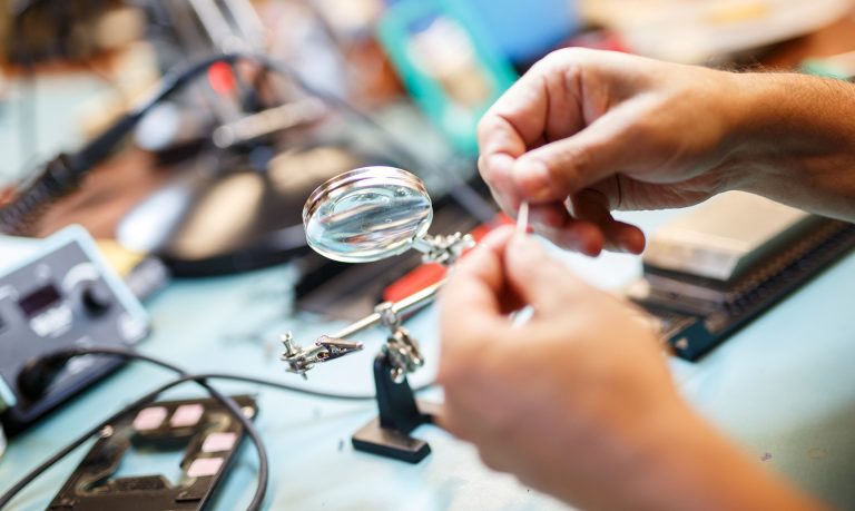 Pictured: Hands holding a small stick, with a table of objects in the background, included a magnifying glass, wire and tools.