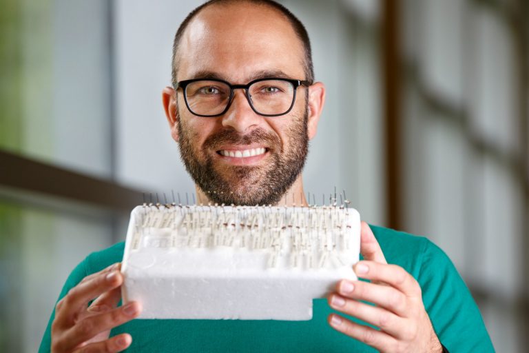 Smiling man with green t-shirt holding piece of white foam with markers and samples.