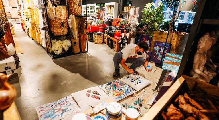 Pictured: A young man crouching on the floor and painting one of four canvases resting on the floor, in a grocery store space.