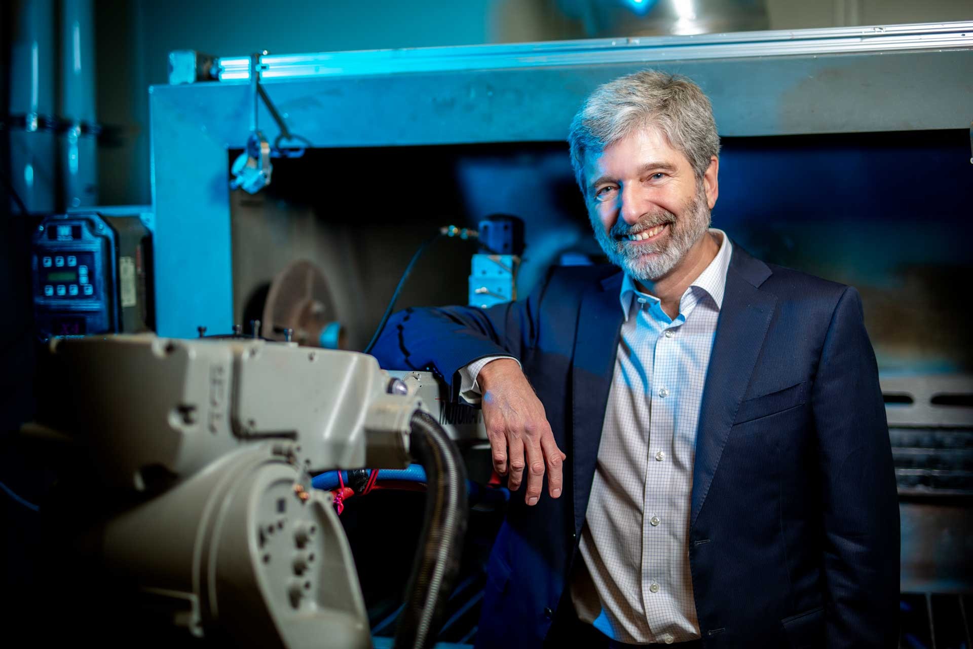 Pictured: A smiling man with short grey hair, a grey beard, in a shirt and jacket and standing next to a piece of machinery.