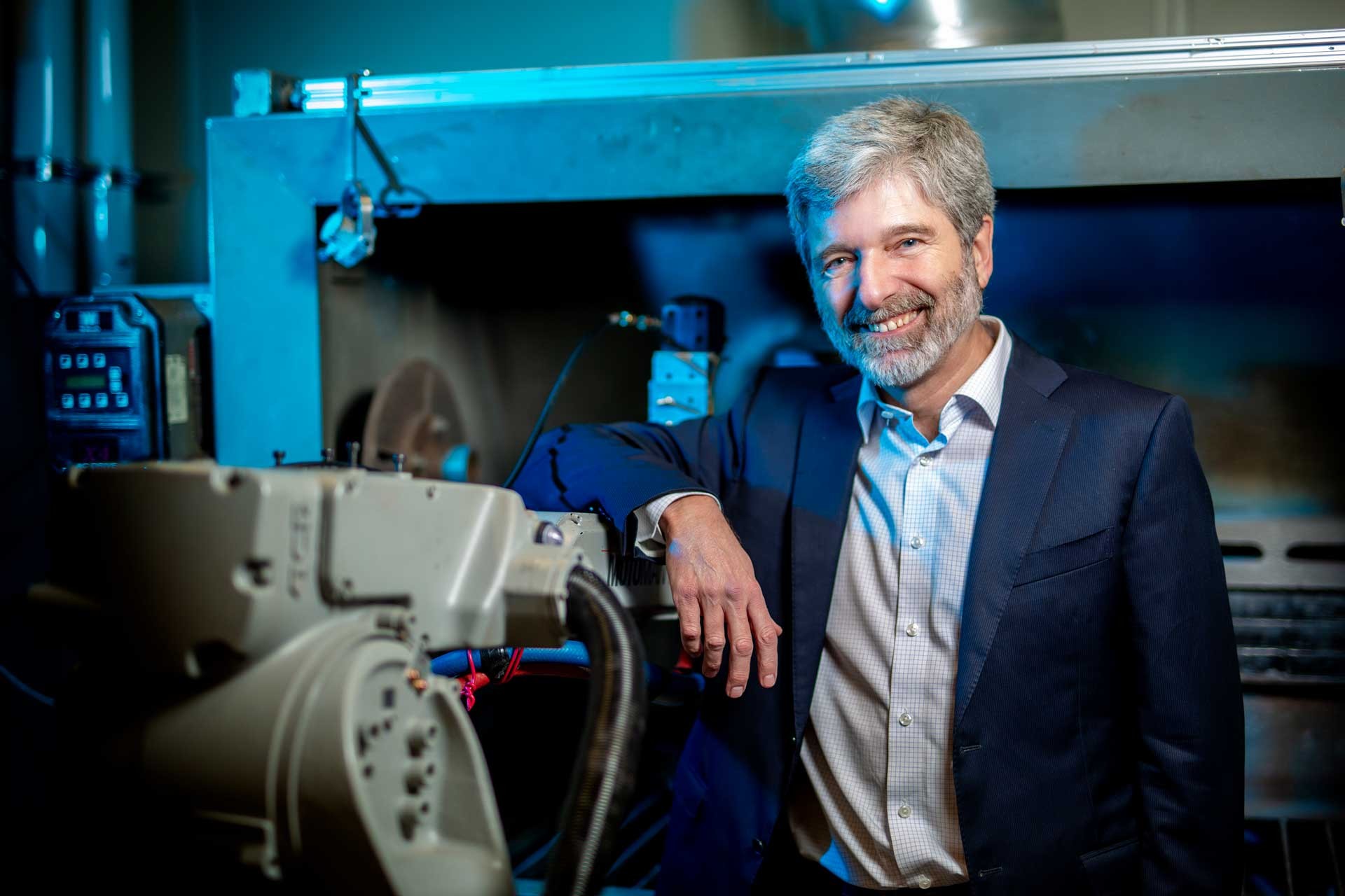 Christian Moreau: “This new approach has the potential to revolutionize our manufacturing processes to produce coatings of exceptional quality.” Pictured: A smiling man with short grey hair, a grey beard, in a shirt and jacket and standing next to a piece of machinery.