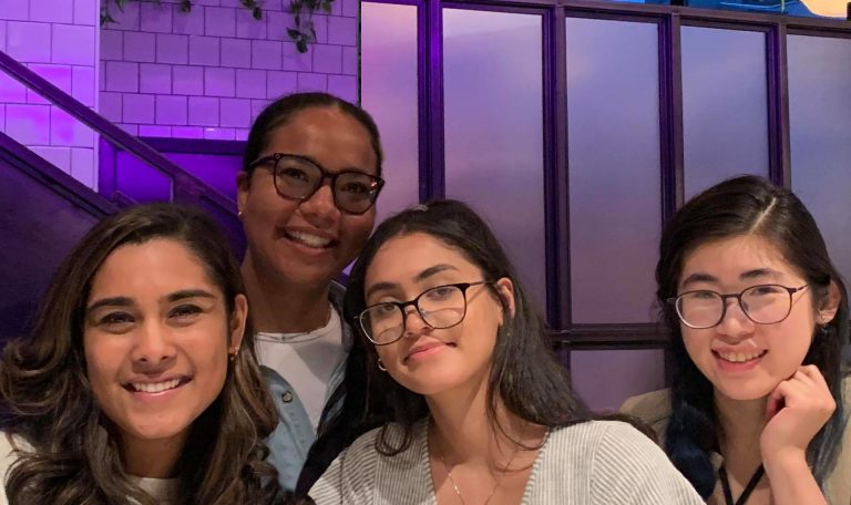 Pictured: Four young women smiling, in front of a purple-lit wall of half brick and half small windows.