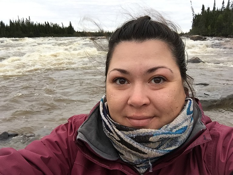 Pictured: Young woman in a winter jacket with long, dark hair tied back and standing in front of a rushing river.