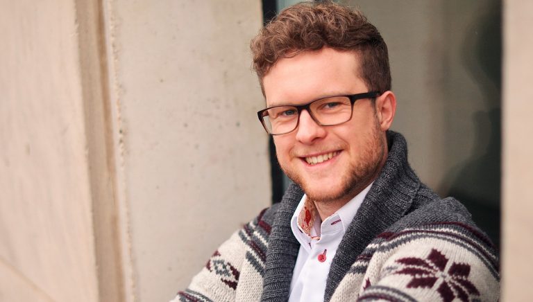 A young smiling man with short, curly hair, glasses and a open-necked sweater.