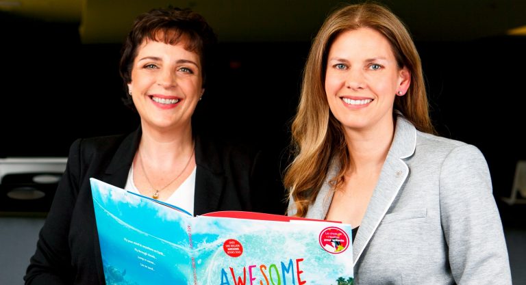 Two women sitting side by side and holding a picture book open between them. On the left, a woman with short, dark hair and on the right, a woman with long blonde hair.