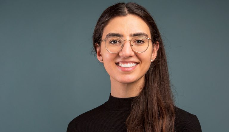 Young smiling woman with long, brown hair, wire-frame glasses and a black turtleneck sweater.